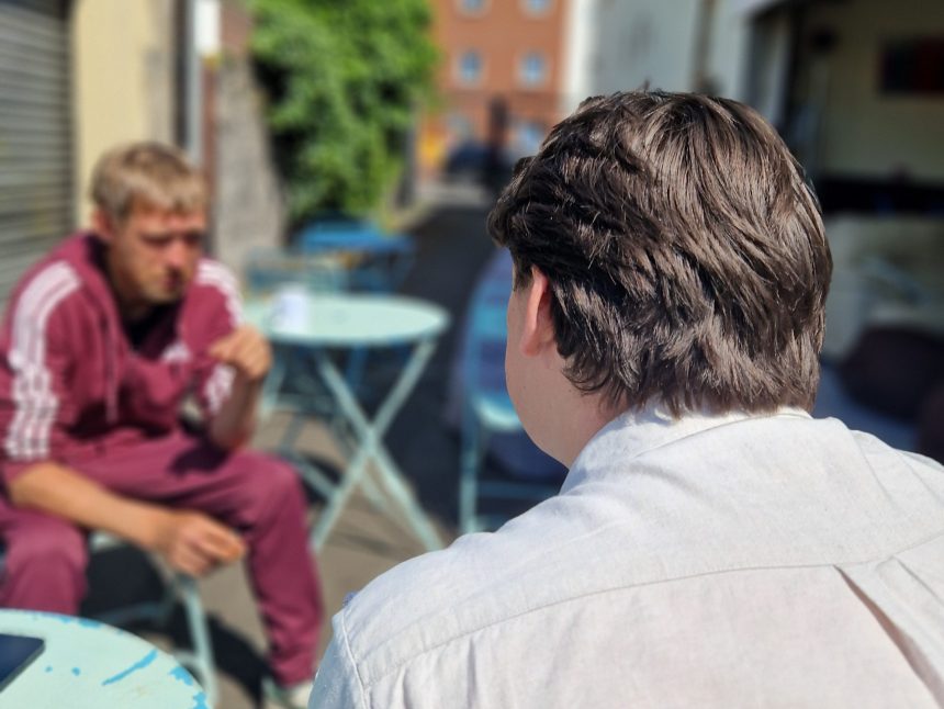 An outreach worker sits talking with a man at an outdoor table at a homelessness drop-in centre.