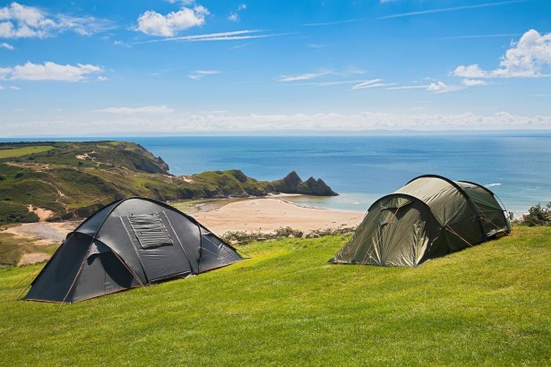 Two tents pitched on a grassy clifftop above Three Cliffs Bay, with the beach, sea and the three limestone peaks in the background under a blue sky.