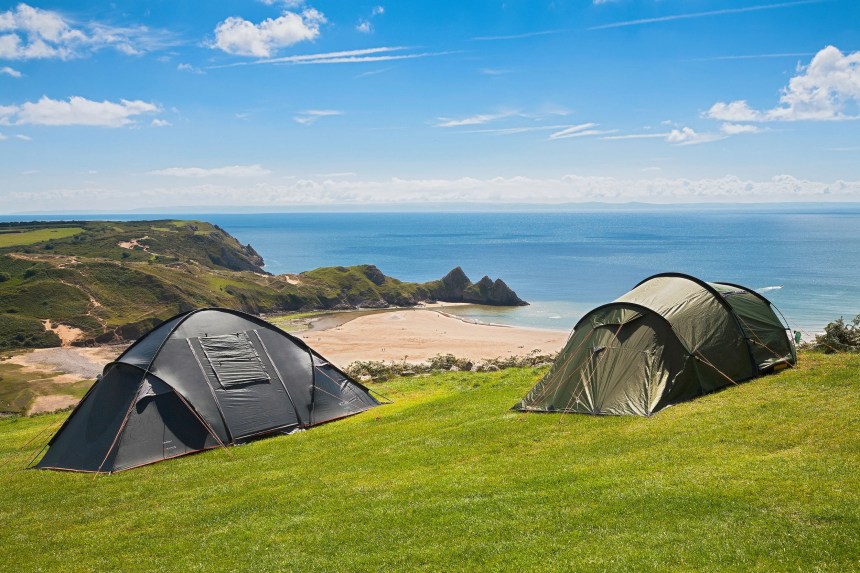 Two tents pitched on a grassy clifftop above Three Cliffs Bay, with the beach, sea and the three limestone peaks in the background under a blue sky.
