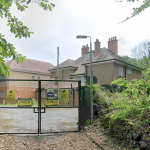 A disused rendered building behind locked metal security gates carrying CCTV and Shield Security warning signs, surrounded by overgrown trees and bushes.