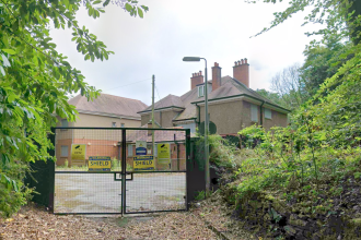 A disused rendered building behind locked metal security gates carrying CCTV and Shield Security warning signs, surrounded by overgrown trees and bushes.