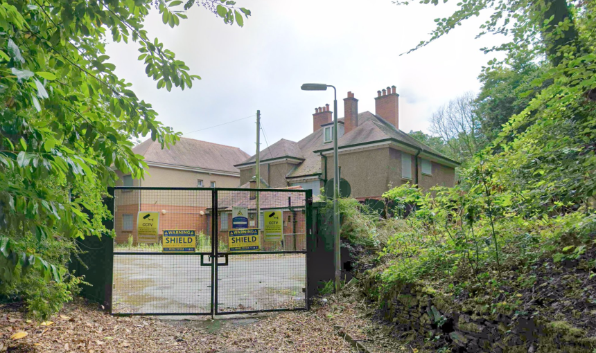A disused rendered building behind locked metal security gates carrying CCTV and Shield Security warning signs, surrounded by overgrown trees and bushes.