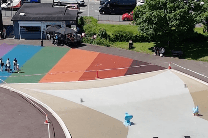 Aerial view of the drained Blackpill Lido, fenced off with hazard tape and traffic cones, the rainbow-coloured surround empty apart from a few people by the kiosk.