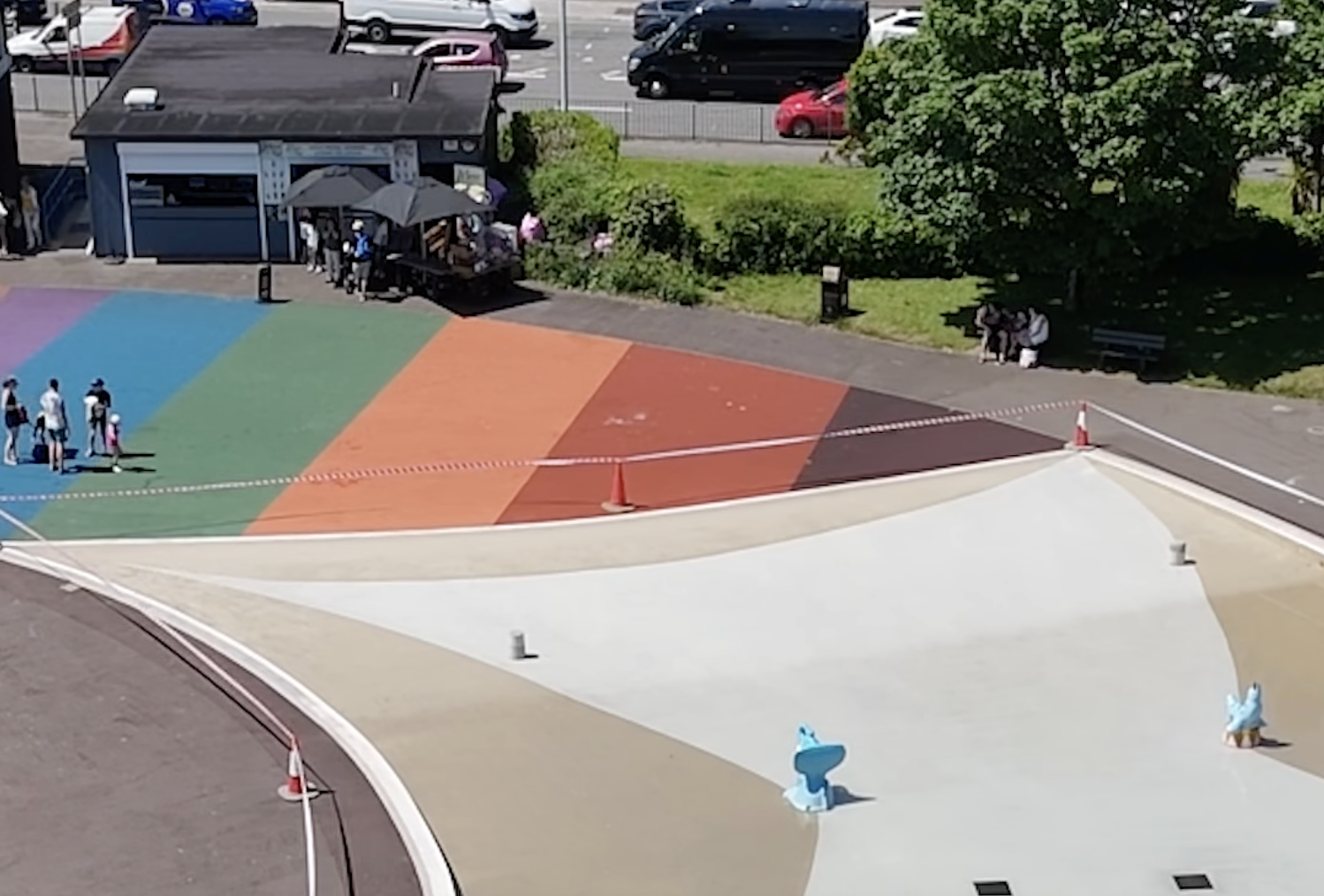 Aerial view of the drained Blackpill Lido, fenced off with hazard tape and traffic cones, the rainbow-coloured surround empty apart from a few people by the kiosk.