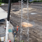 A worker in orange overalls on a raised platform cutting into a steel floodlight tower with sparks flying, above the dug-up pitch and empty terraces at St Helen's, Swansea.