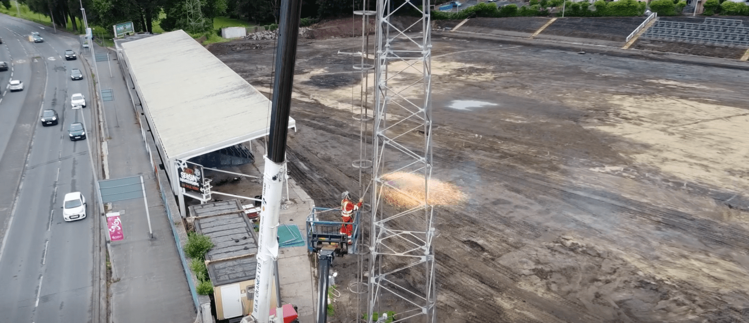 A worker in orange overalls on a raised platform cutting into a steel floodlight tower with sparks flying, above the dug-up pitch and empty terraces at St Helen's, Swansea.