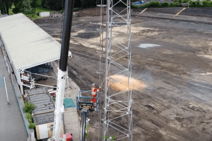 A worker in orange overalls on a raised platform cutting into a steel floodlight tower with sparks flying, above the dug-up pitch and empty terraces at St Helen's, Swansea.
