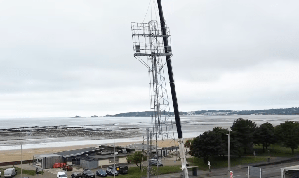 A steel floodlight pylon suspended by a crane at St Helen's stadium, Swansea, with the sweep of Swansea Bay and Mumbles headland behind.