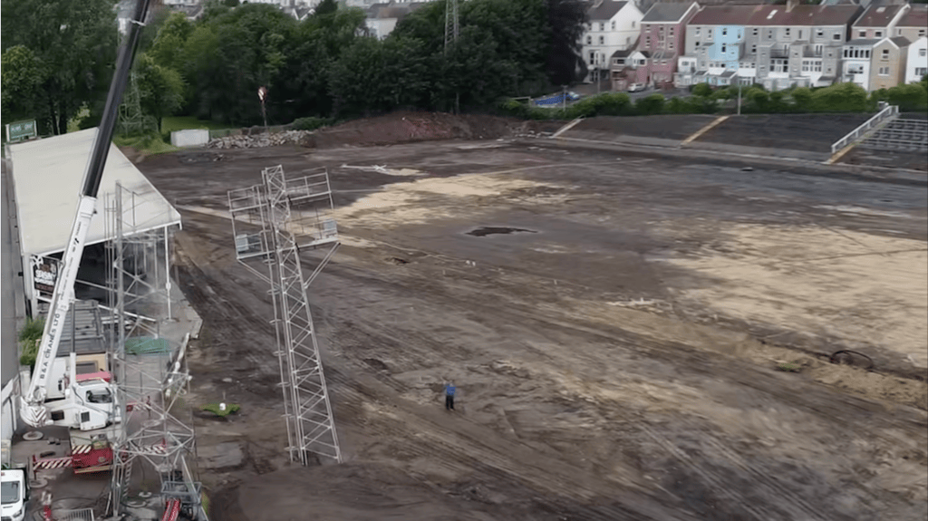 An aerial view of St Helen's stadium in Swansea with the playing surface dug up to bare earth, a steel floodlight pylon and a mobile crane on site, terraced houses behind.
