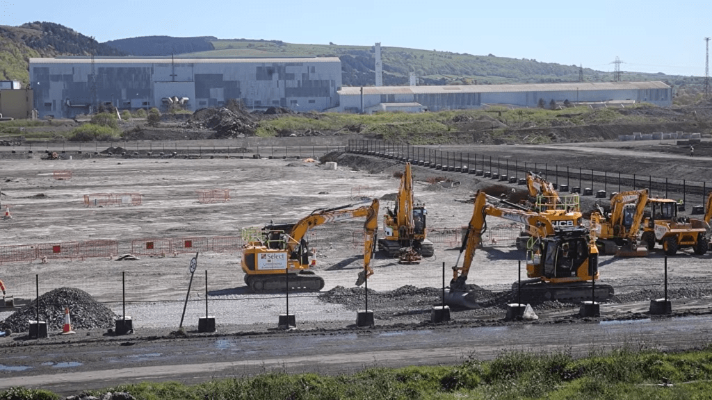 Several yellow excavators working across a large cleared construction site, with steelworks buildings on the hillside behind.