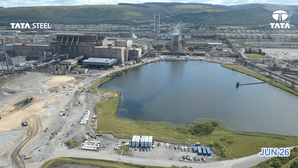 An aerial view of a large dark lagoon beside steelworks buildings, with green hills behind.