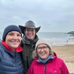 Three women smiling at a Gower beach on an overcast day, with the sea and a bilingual beach sign behind them.