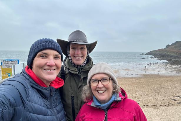 Three women smiling at a Gower beach on an overcast day, with the sea and a bilingual beach sign behind them.