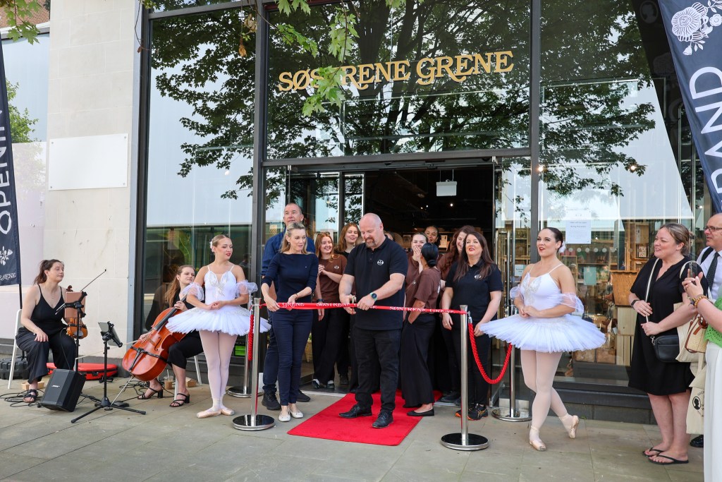 A man cuts a red ribbon outside the new store as staff look on and ballet dancers pose on either side.