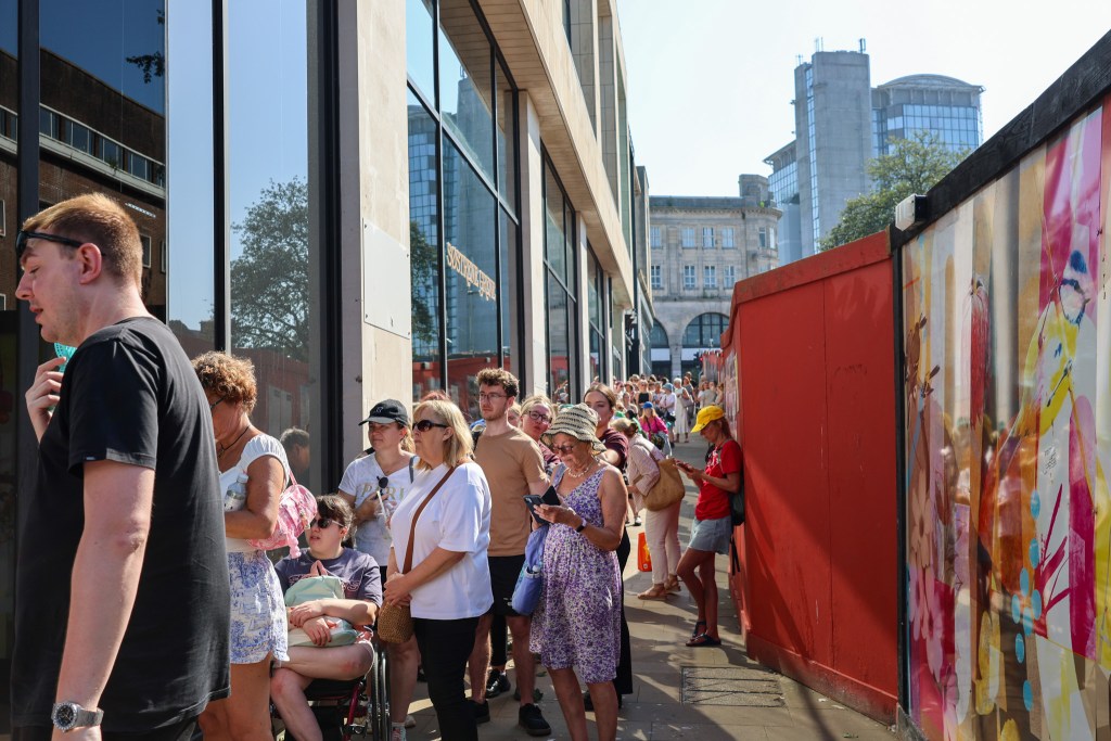 A long queue of people along the store's glass frontage in bright sunshine, with city-centre buildings behind.