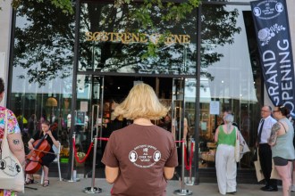 The Søstrene Grene storefront under its gold sign, with a "Grand Opening" banner, a cellist and shoppers gathering by the entrance.