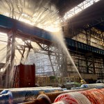 A firefighter directs a jet of water high into the steel roof structure of the Tata steelworks mill, with sunlight streaming through gaps in the damaged roof above stacked steel rolls.