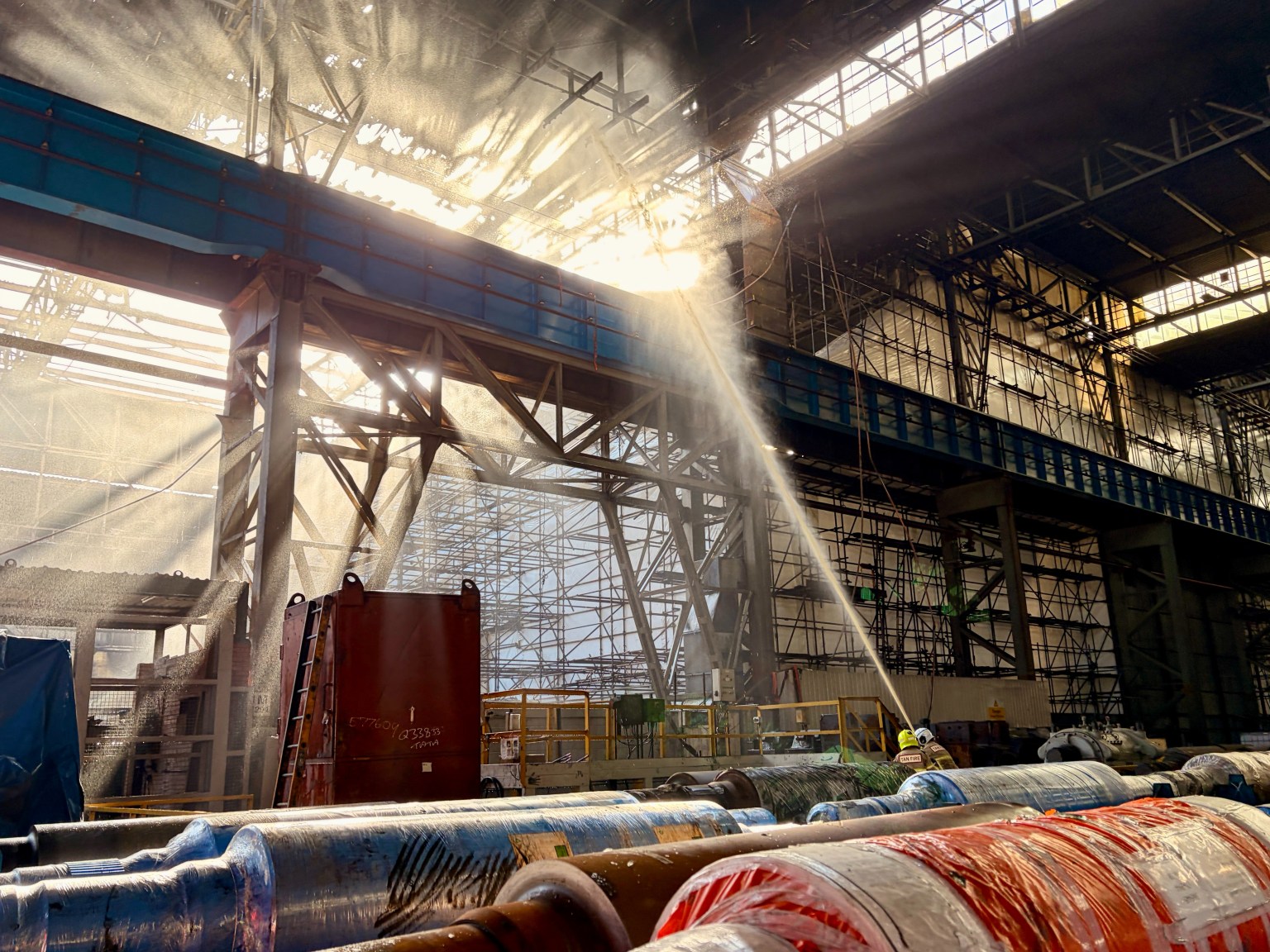 A firefighter directs a jet of water high into the steel roof structure of the Tata steelworks mill, with sunlight streaming through gaps in the damaged roof above stacked steel rolls.