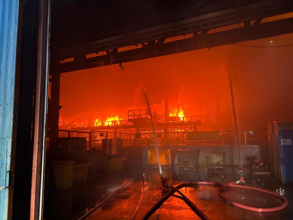 Orange flames and thick smoke fill the inside of an industrial building as two jets of water are directed at the fire.