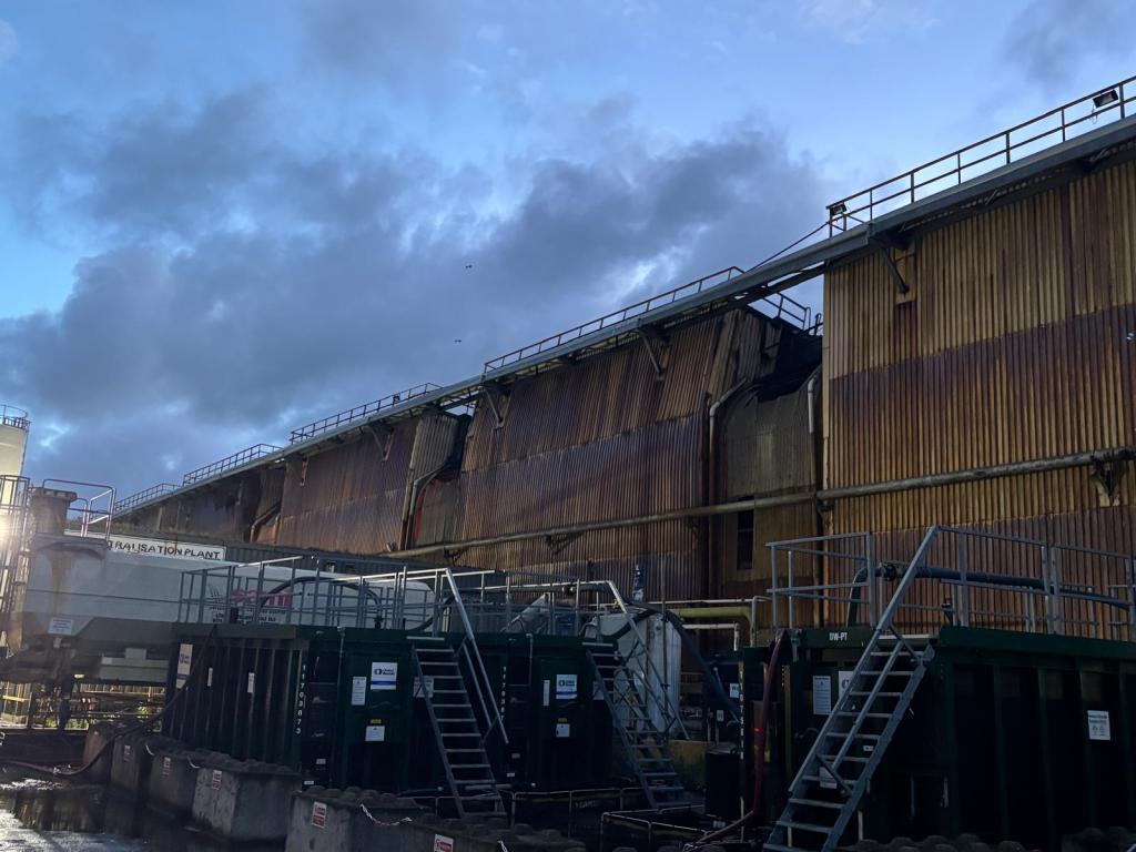 Scorched and buckled metal cladding on a large steelworks building, seen in daylight under a cloudy sky.