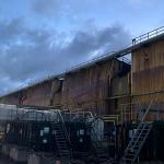 Scorched and buckled metal cladding on a large steelworks building, seen in daylight under a cloudy sky.