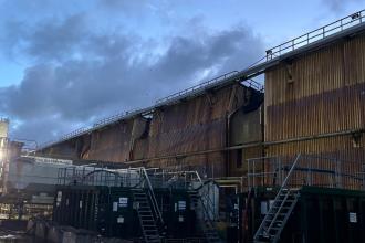 Scorched and buckled metal cladding on a large steelworks building, seen in daylight under a cloudy sky.