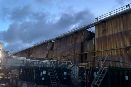 Scorched and buckled metal cladding on a large steelworks building, seen in daylight under a cloudy sky.