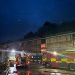 A fire engine and firefighter outside a steelworks building at night with dark smoke overhead.