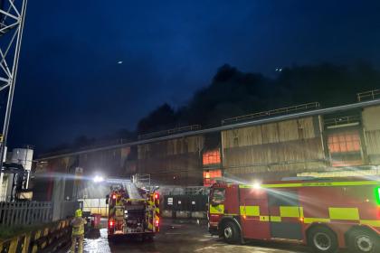A fire engine and firefighter outside a steelworks building at night with dark smoke overhead.