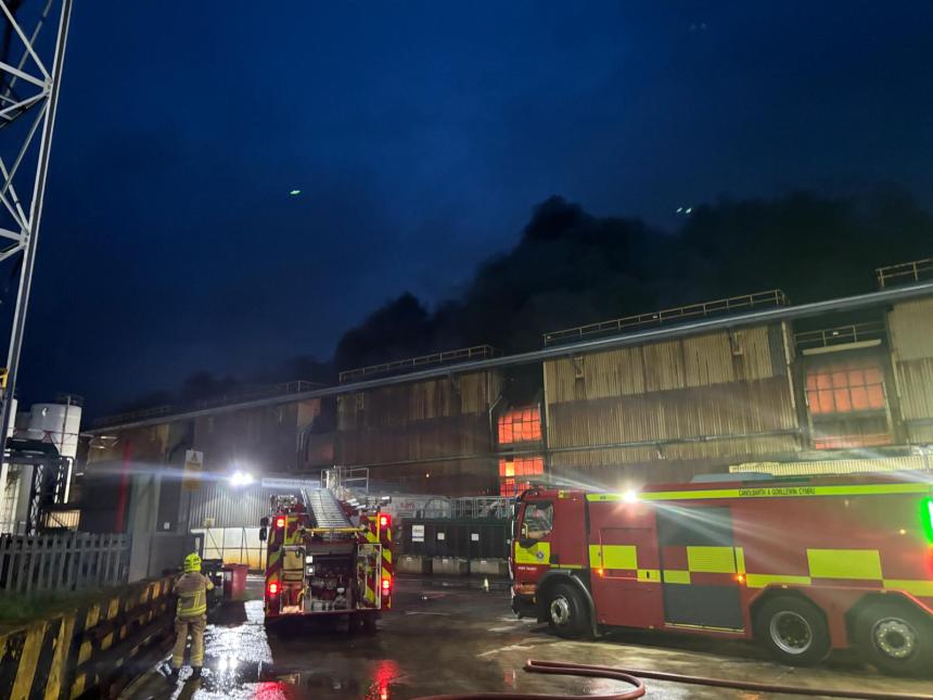 A fire engine and firefighter outside a steelworks building at night with dark smoke overhead.