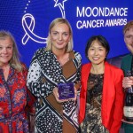 Three women and a man on stage at the Moondance Cancer Awards, one holding the Better Patient Experience award and another holding a bottle of sparkling wine, in front of a blue screen with the awards logo