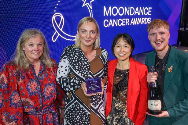 Three women and a man on stage at the Moondance Cancer Awards, one holding the Better Patient Experience award and another holding a bottle of sparkling wine, in front of a blue screen with the awards logo