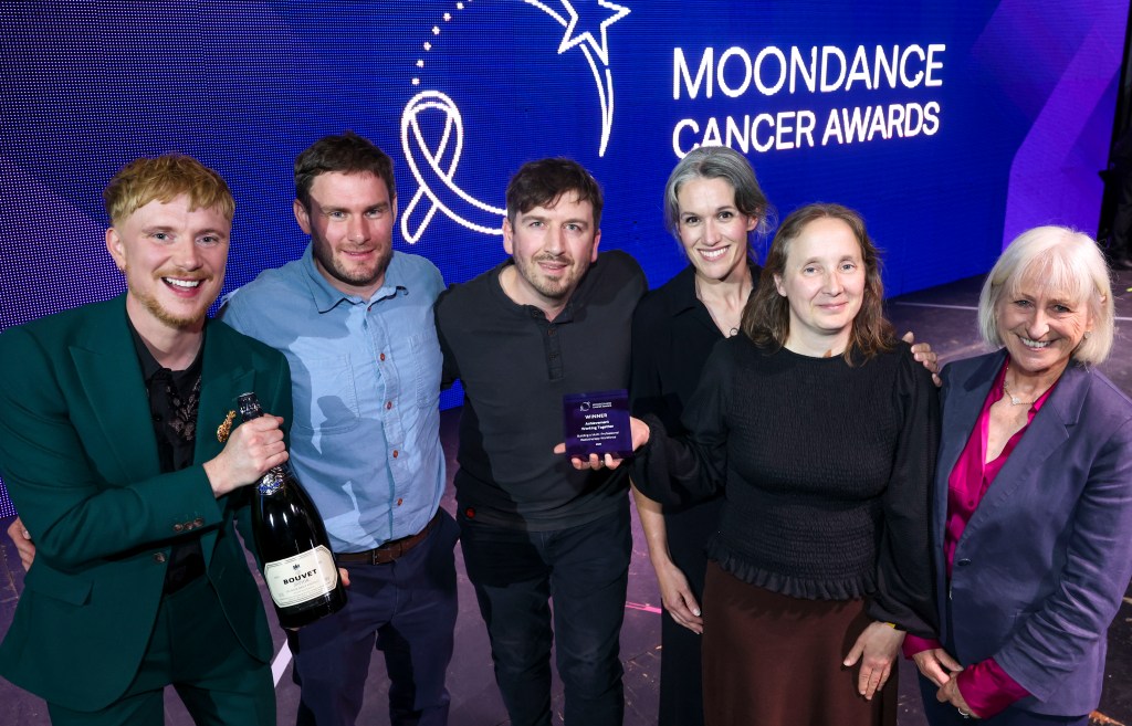 Six people on stage at the Moondance Cancer Awards, with a man at the centre holding the Working Together award and another holding a bottle of sparkling wine, in front of a blue awards screen