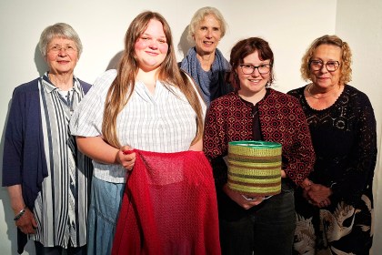 Five women standing against a white wall, two holding textile pieces — a red shawl and a striped woven lampshade