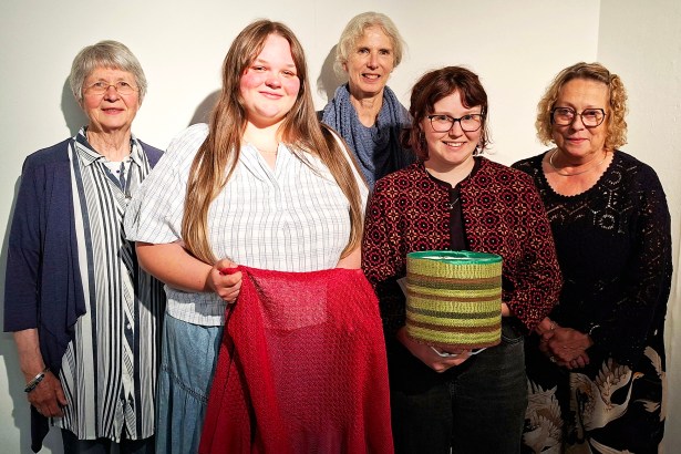 Five women standing against a white wall, two holding textile pieces — a red shawl and a striped woven lampshade