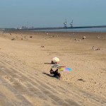 A wide view of Aberavon Beach in the sunshine, with people on the sand and the docks cranes visible on the horizon.
