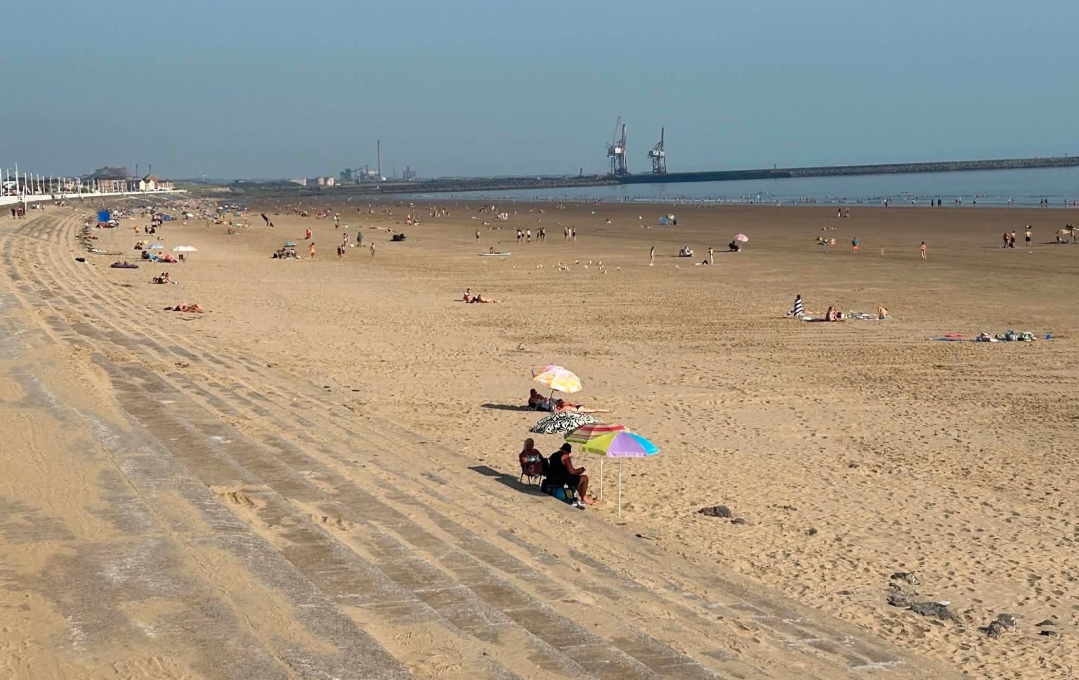 A wide view of Aberavon Beach in the sunshine, with people on the sand and the docks cranes visible on the horizon.