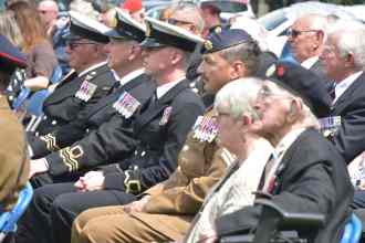 A row of seated veterans and serving personnel in naval and army uniforms with medals, watching the ceremony in the sunshine.