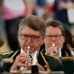 Musicians in green and gold military-style uniforms playing brass instruments at an outdoor Armed Forces Day event, with a crowd behind them.