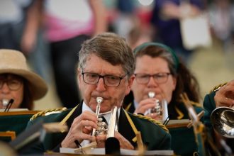 Musicians in green and gold military-style uniforms playing brass instruments at an outdoor Armed Forces Day event, with a crowd behind them.