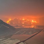 Flames and thick smoke rising from a large fire at a waste recycling site at night, with a fire hose running across the foreground.
