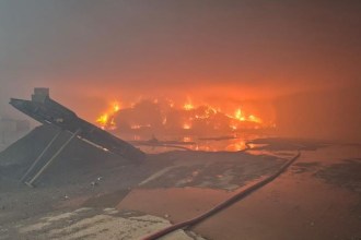 Flames and thick smoke rising from a large fire at a waste recycling site at night, with a fire hose running across the foreground.