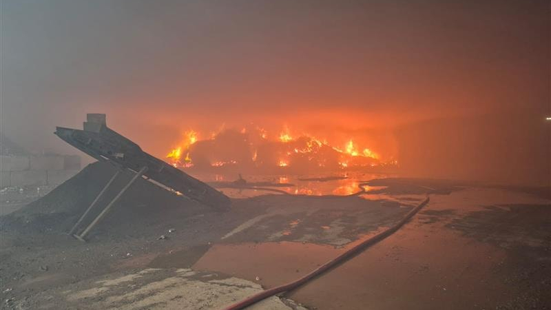 Flames and thick smoke rising from a large fire at a waste recycling site at night, with a fire hose running across the foreground.