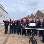 A male voice choir in black, waving and smiling on a dockside in front of a cruise ship.