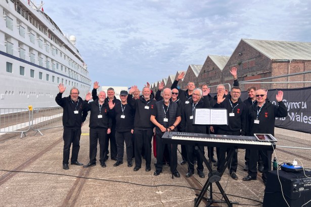 A male voice choir in black, waving and smiling on a dockside in front of a cruise ship.