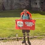 A man with a backpack holding a Brain Tumour Charity banner in front of Chepstow Castle.