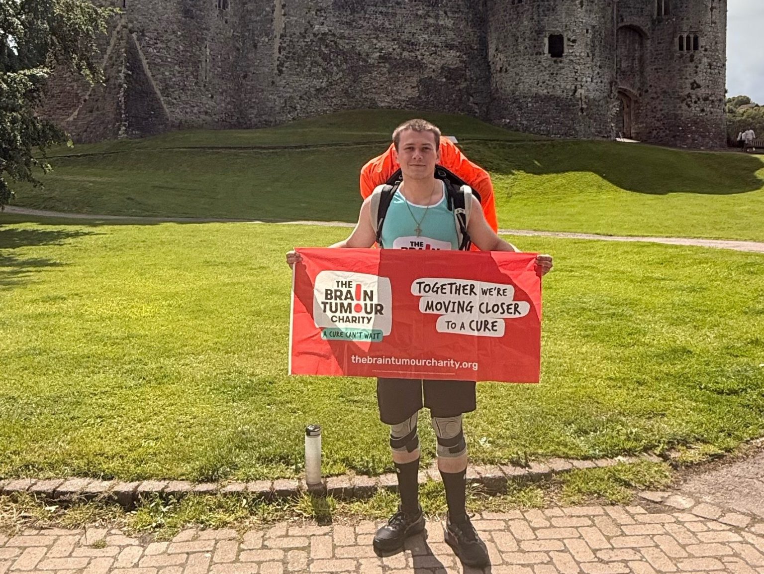 A man with a backpack holding a Brain Tumour Charity banner in front of Chepstow Castle.