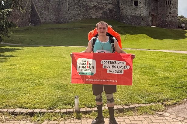 A man with a backpack holding a Brain Tumour Charity banner in front of Chepstow Castle.