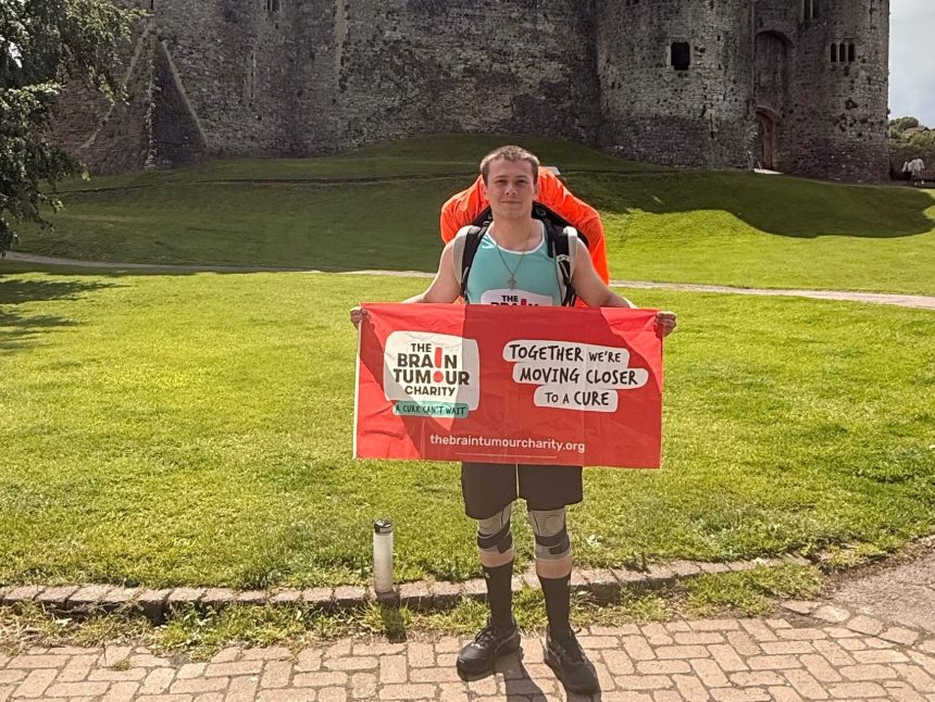 A man with a backpack holding a Brain Tumour Charity banner in front of Chepstow Castle.
