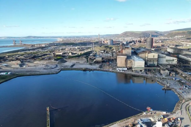 Aerial view of the Port Talbot steelworks, with a large water reservoir in the foreground, blast furnaces and chimneys beyond, and Swansea Bay in the distance.